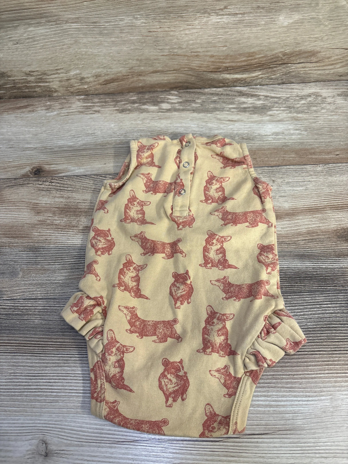 Flat lay of tan Kate Quinn Korgi bodysuit with rust corgi print, snap neck, and ruffled leg openings on light wood background