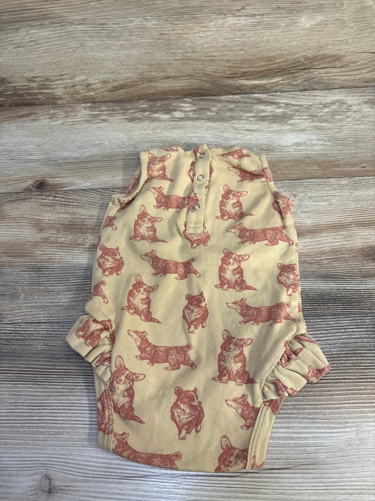 Flat lay of Kate Quinn Korgi Bodysuit in tan sz 12-18m showcasing allover corgi print and button placket on wood plank floor
