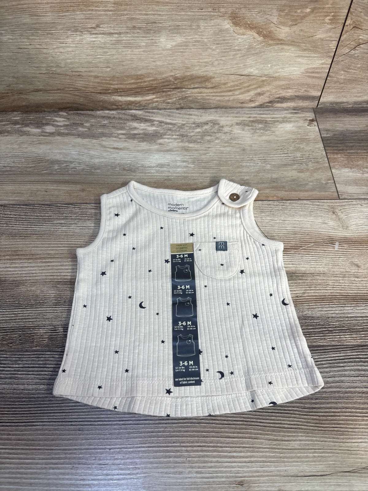 Front view of beige Modern Moments ribbed tank top with star and moon print, shoulder button, and wood-look background