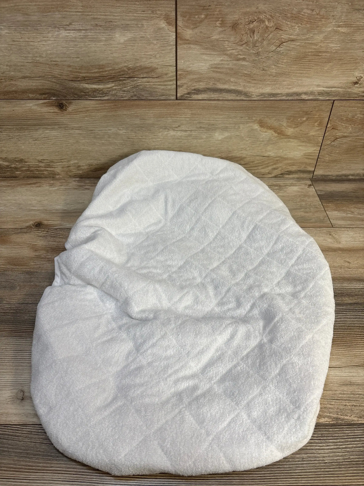 Top-down view of Biloban bassinet mattress pad cover showcasing white quilted surface and fitted shape on wood floor