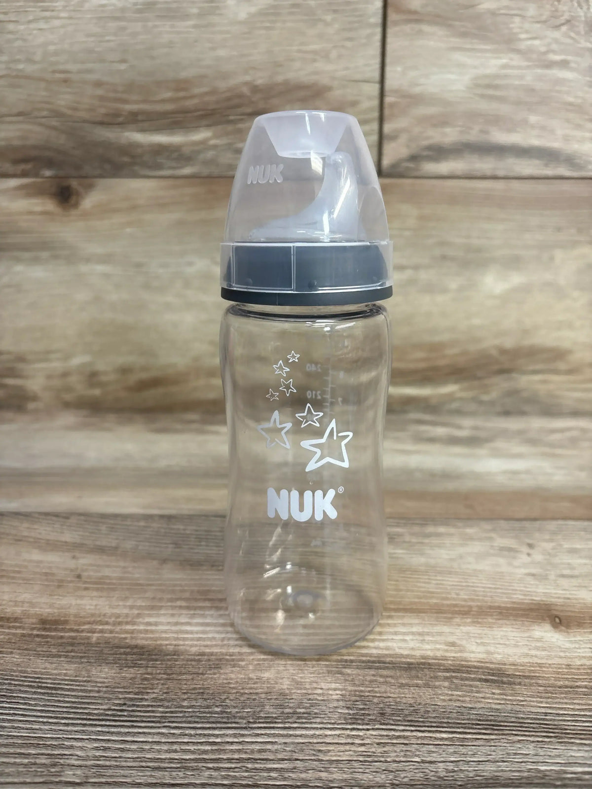 Front view of NUK 10oz sippy cup showcasing clear bottle with white star graphics, gray spill-proof spout and measurement markings on wood background