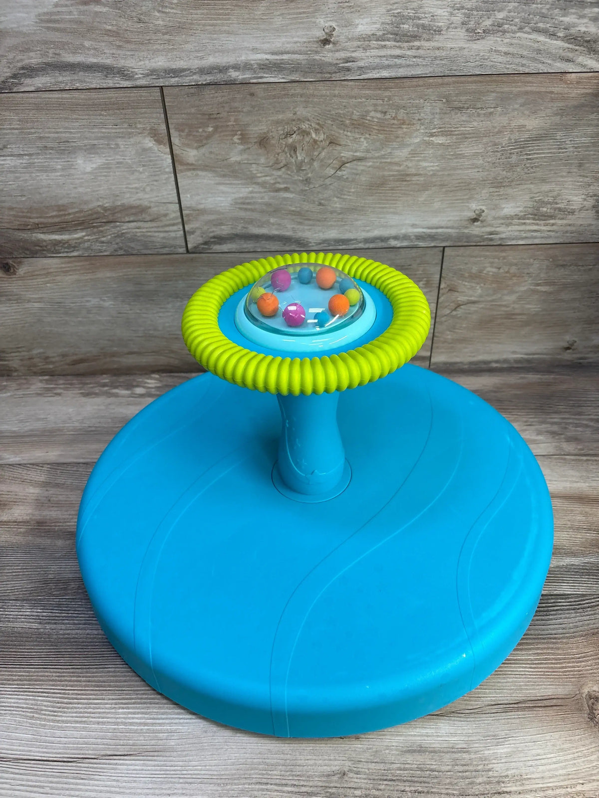 Three-quarter view of B Toys Twirly Time Spinning Activity Toy showcasing teal base green ribbed grip and clear dome with colorful balls on wood plank background