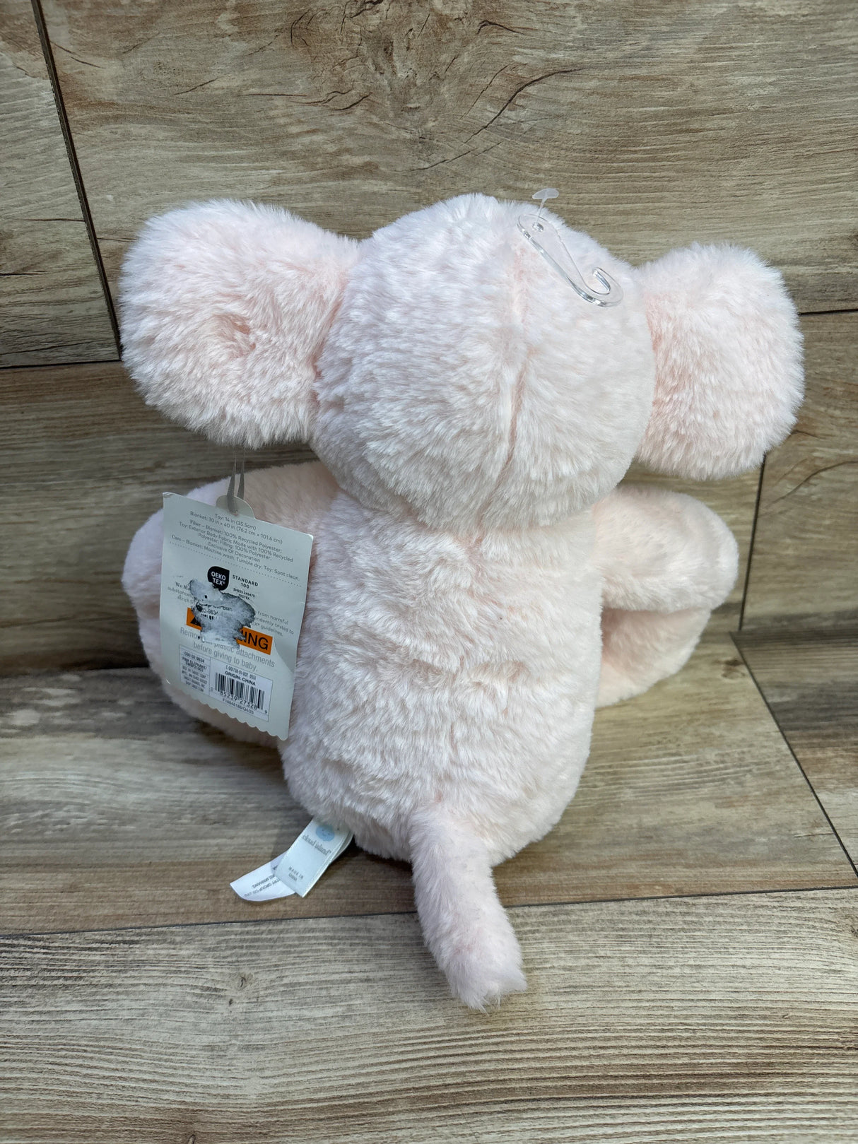 Back view of Cloud Island pink elephant plush showing soft fuzzy fur, small tail, and retail tags on a wood floor background
