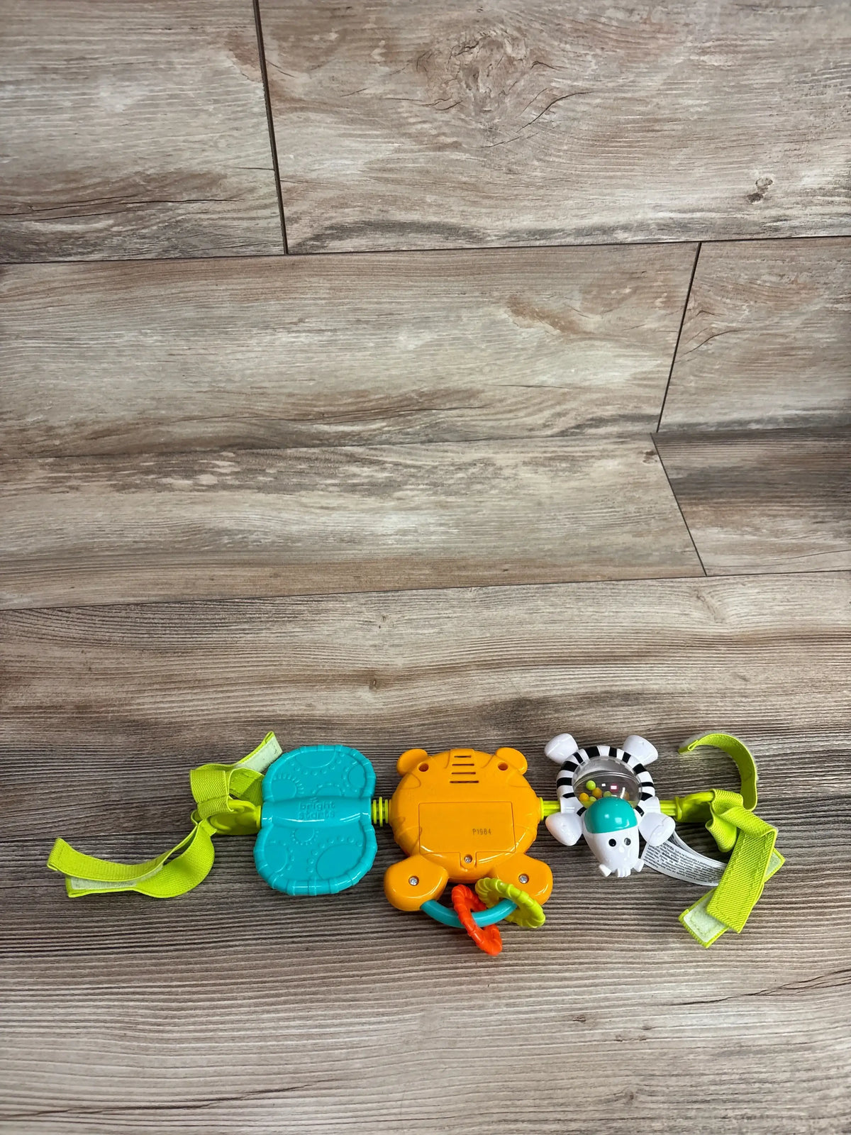 Top view of Bright Starts take along carrier toy bar showcasing teal teether orange tiger rattle and zebra clip toy with green straps on wood plank floor