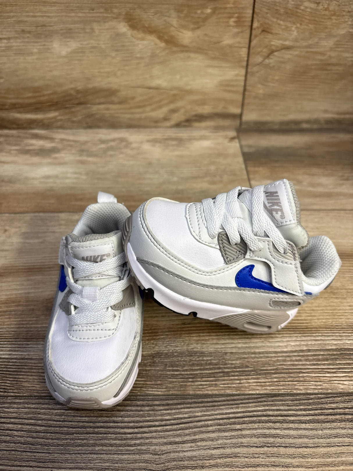 Front and side view of Nike Air Max 90 EasyOn toddler sneakers showing blue Swoosh and hook-and-loop strap on wood background