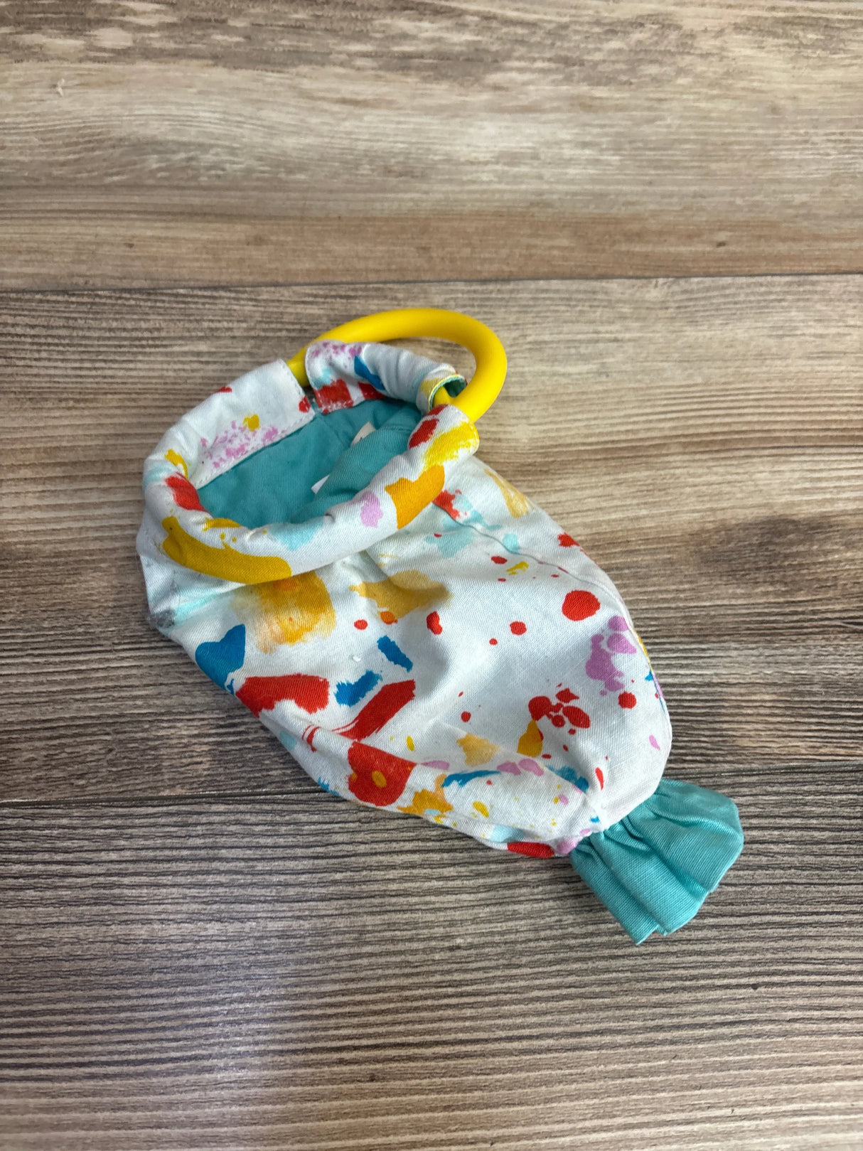 Top-down view of Lovevery Organic Cotton sensory pouch with yellow teething ring and multicolor splatter fabric on wood surface