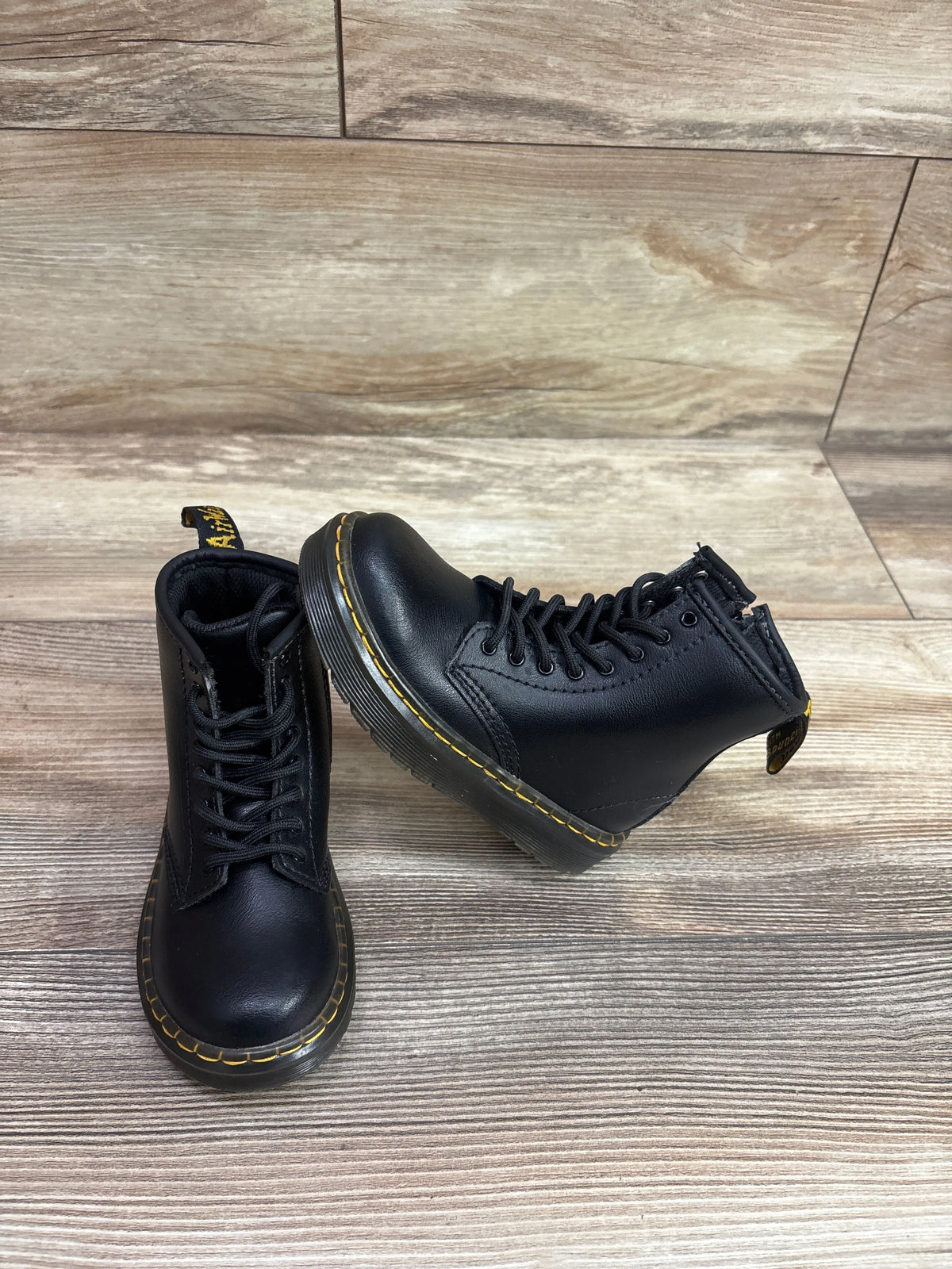 Pair of black Dr. Martens kid's boots with yellow stitching on a wooden background.