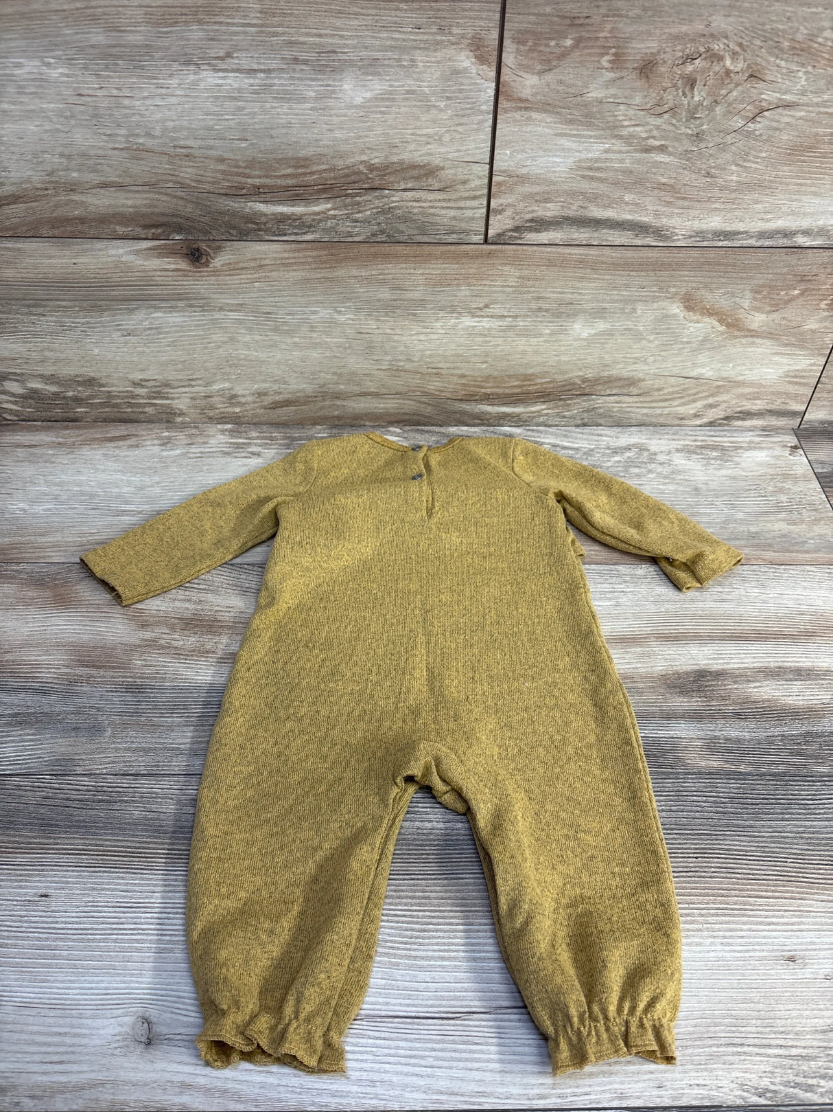 Flat lay of 7 For All Mankind yellow ruffle coverall size 3-6 months showing long sleeves, ruffled ankle cuffs, and back snap closure on wood plank background