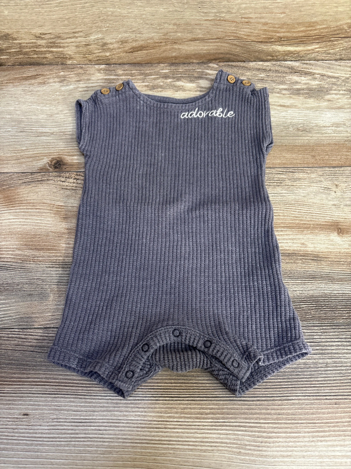 Flat lay of Grayson Mini Knit romper in grey ribbed knit with adorable embroidery, wooden shoulder buttons and snap crotch on wood background