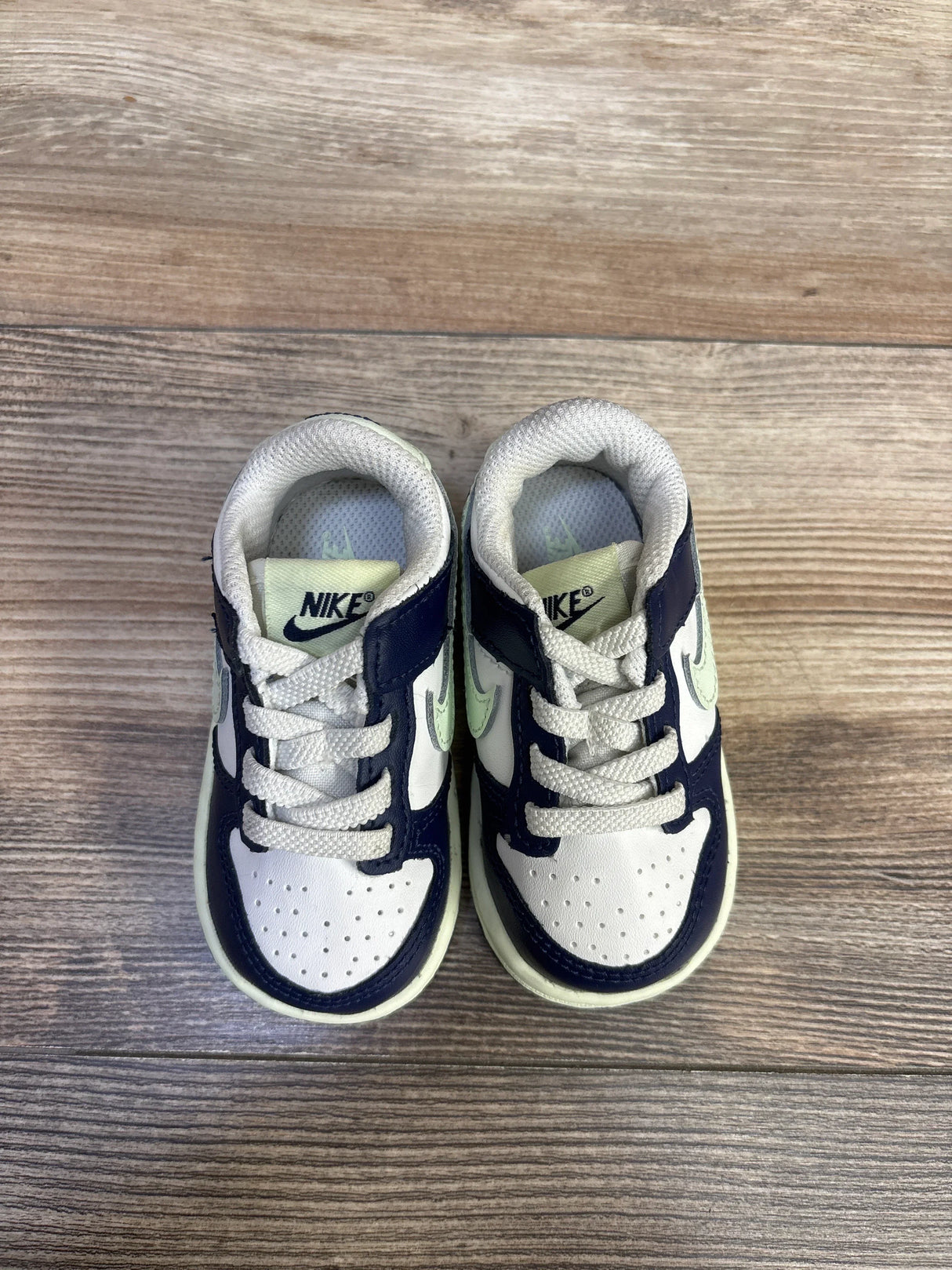 Top view of toddler Nike Dunk Low sneakers in white, midnight navy, volt tint showing laces, perforated toe on wood floor