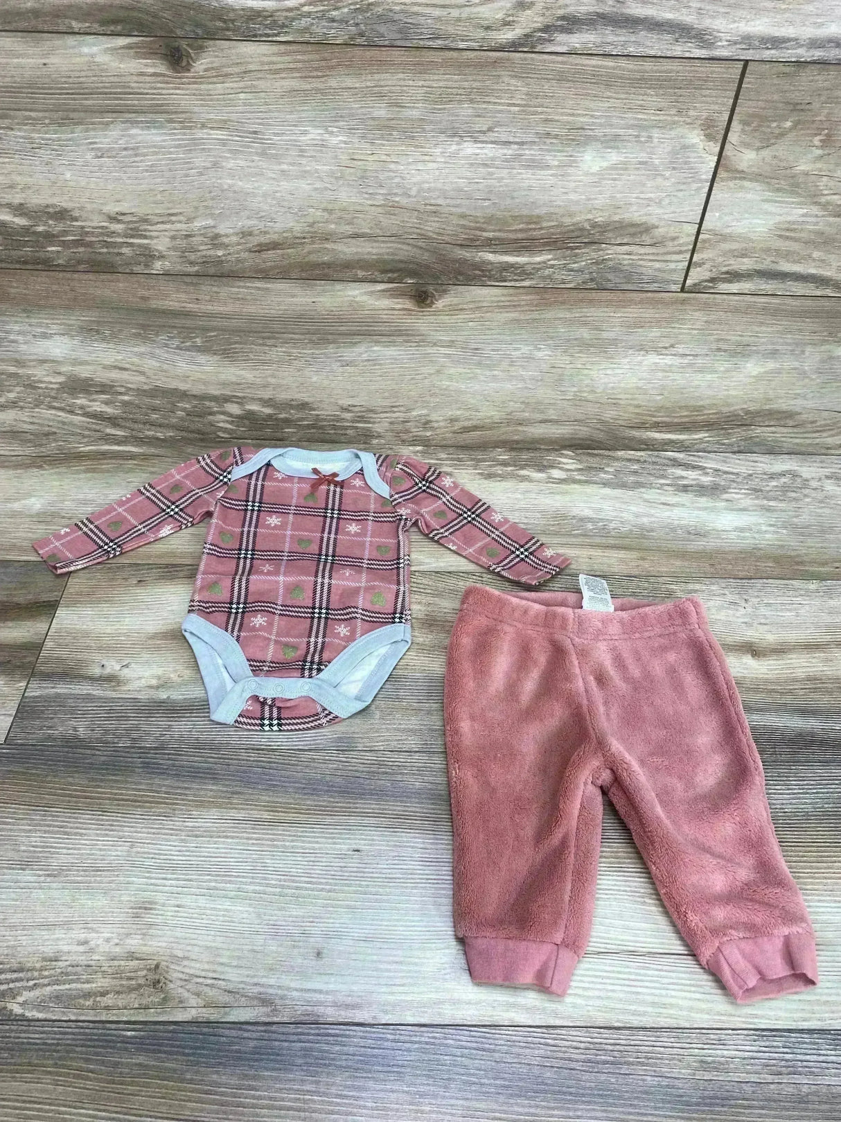Flat lay of Rudolph pink long-sleeve bodysuit with plaid reindeer print and pink fleece pants on wood floor