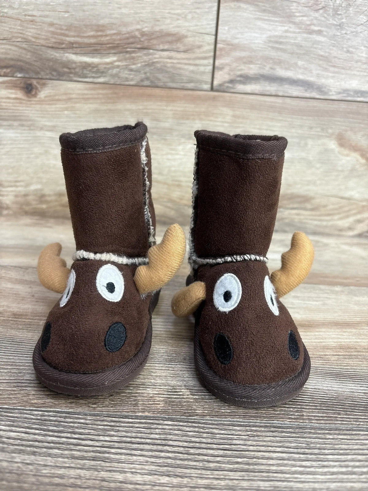 Front view of Lazy One moose slipper boots in brown showcasing 3D antlers and embroidered face with sherpa lining on wood plank floor