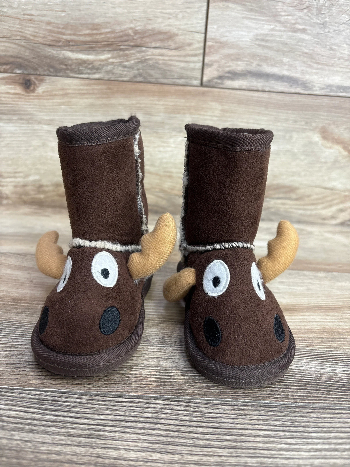 Front view of Lazy One moose slipper boots in brown showcasing 3D antlers and embroidered face with sherpa lining on wood plank floor