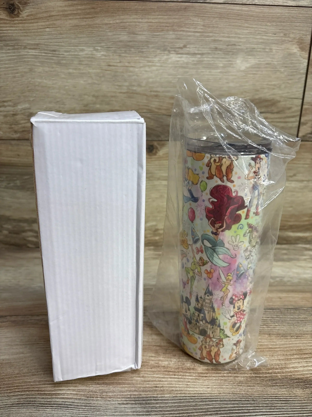 Disney-themed insulated tumbler with characters like Mickey, Minnie, Ariel, and castle, next to white box