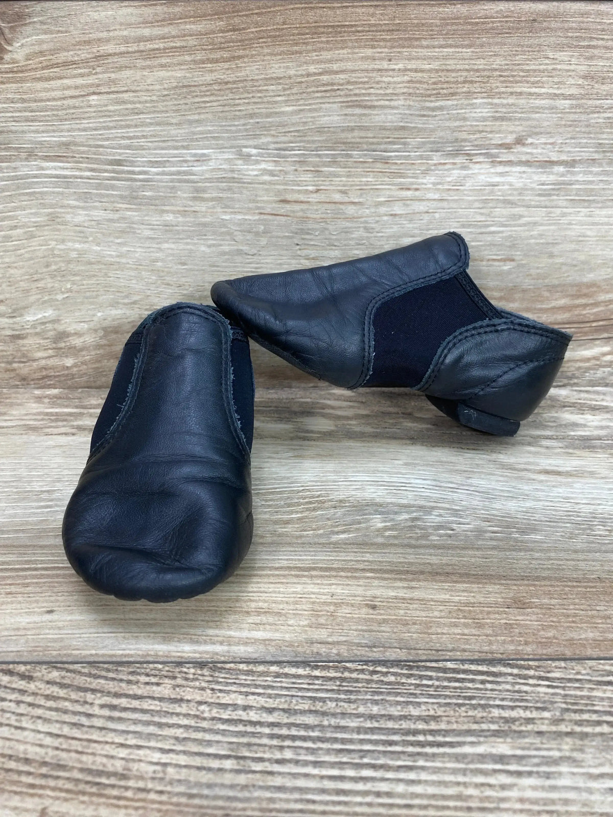 Front-left view of Capezio E-Series black leather jazz slip-on shoes showing elastic side panels and split sole on light wood background