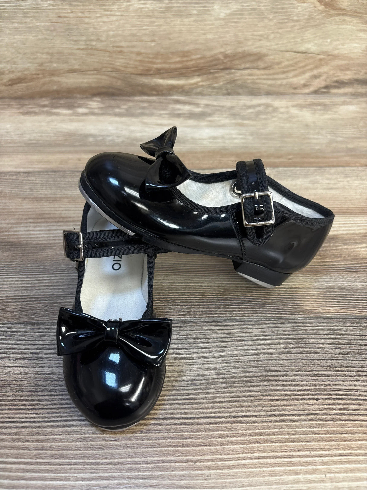 Black patent leather girls' dress shoes with bows and buckle straps on wooden floor