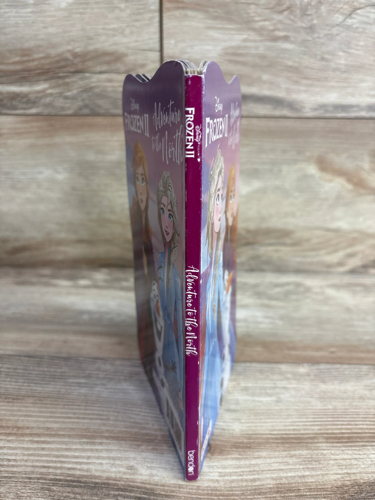 Spine view of Frozen II Adventure to the North board book with scalloped top edge and covers showing Elsa, Anna, and Olaf on wood