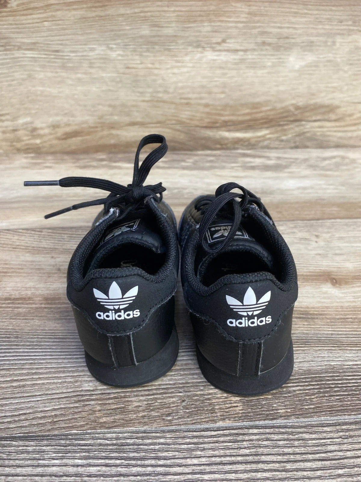 Black Adidas sneakers with white trefoil logo on heels, placed on wooden floor