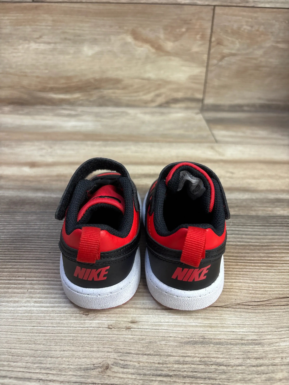 Rear view of Nike Court Borough Recraft TD sneakers showing red and black uppers, red pull tabs, and white soles on wood