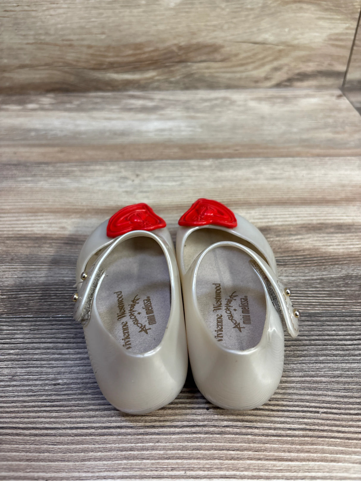 Top view of Vivienne Westwood x Mini Melissa toddler Mary Jane shoes with cream glossy jelly, red orb toe accents, and gold-button straps on a wood tabletop