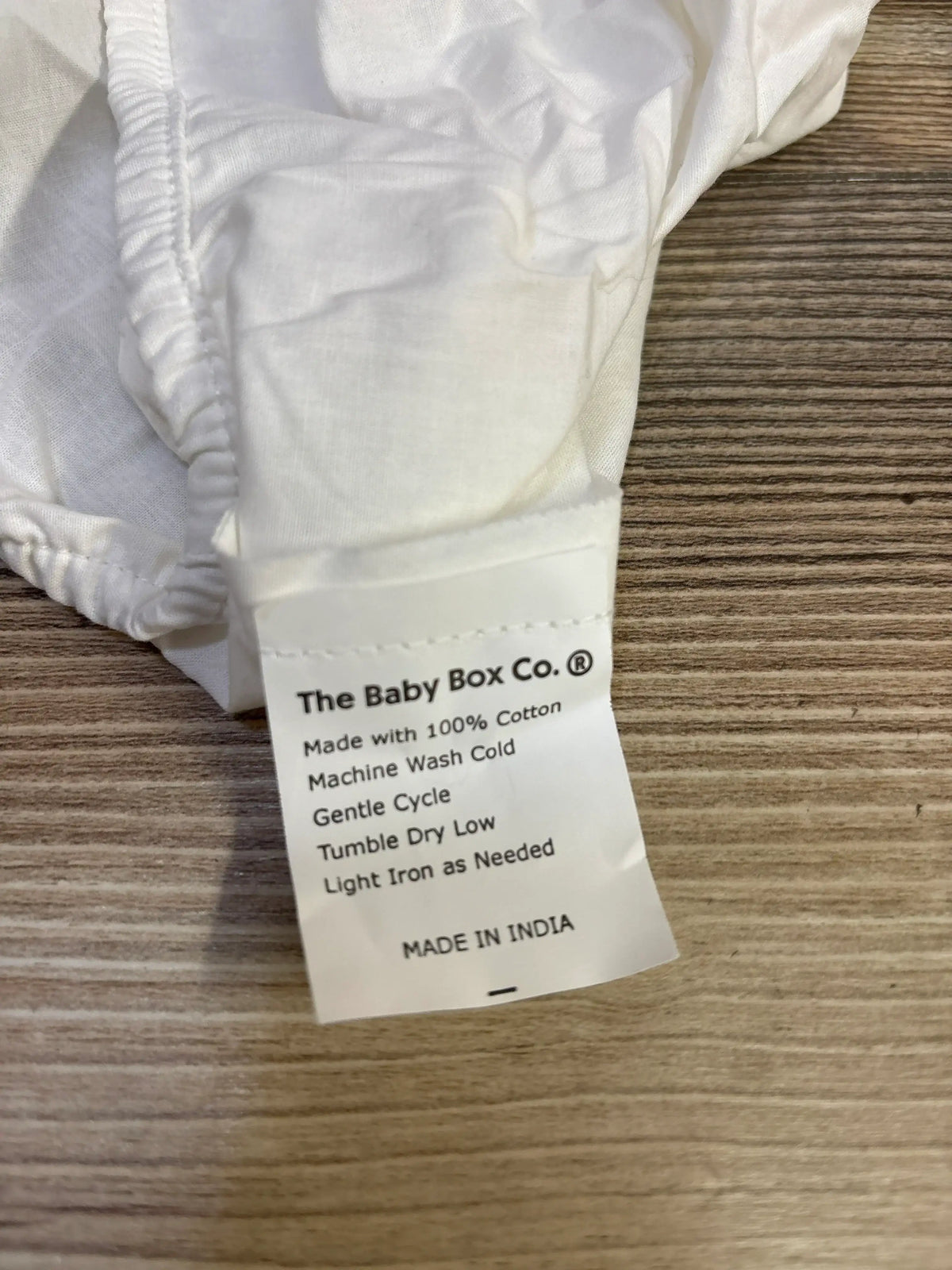 Close-up of The Baby Box Co fitted bassinet sheet showing care label stating 100 percent cotton and Made in India on wood surface