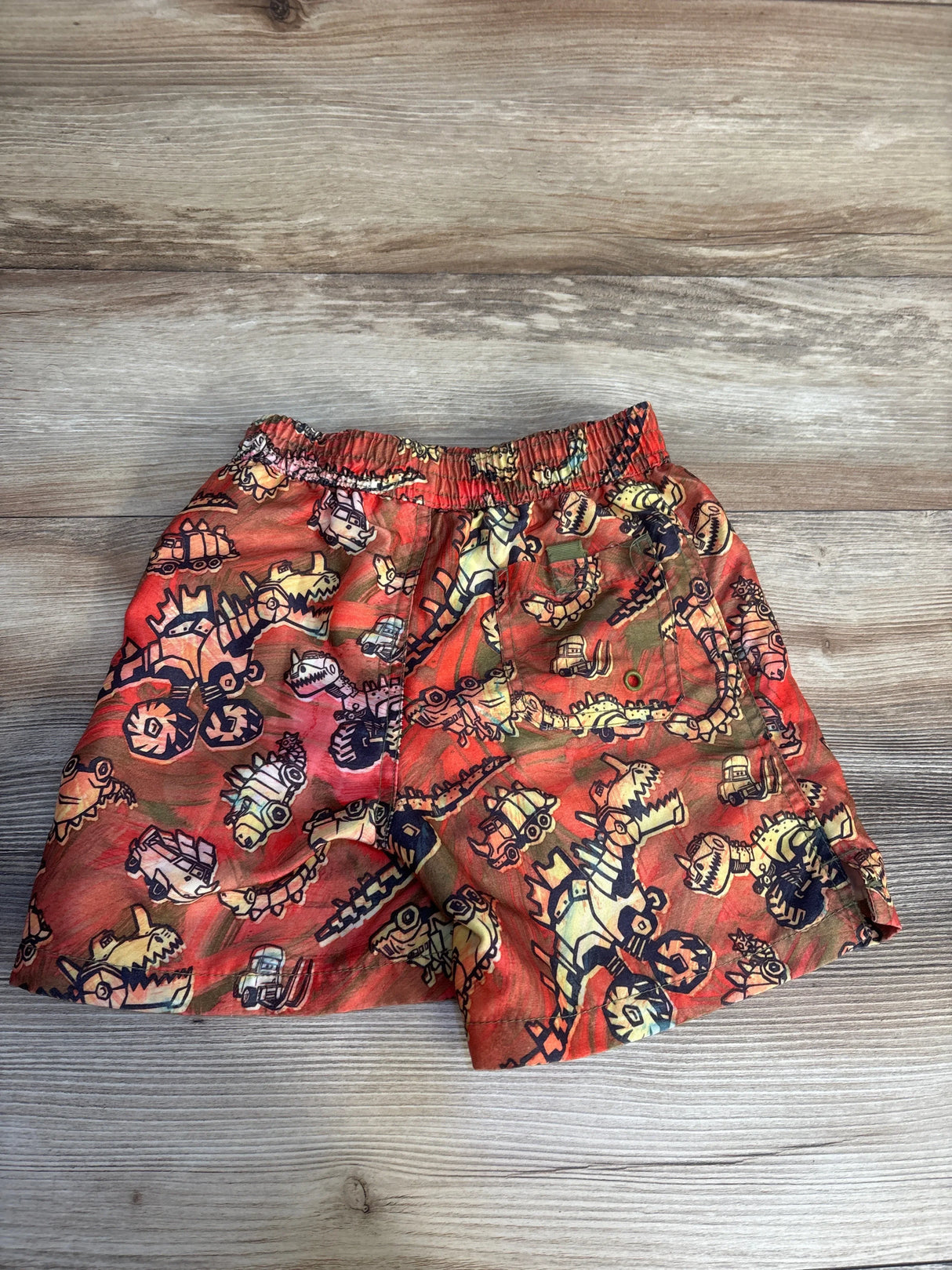 Red toddler shorts with robot dinosaur print, laid flat on wood background