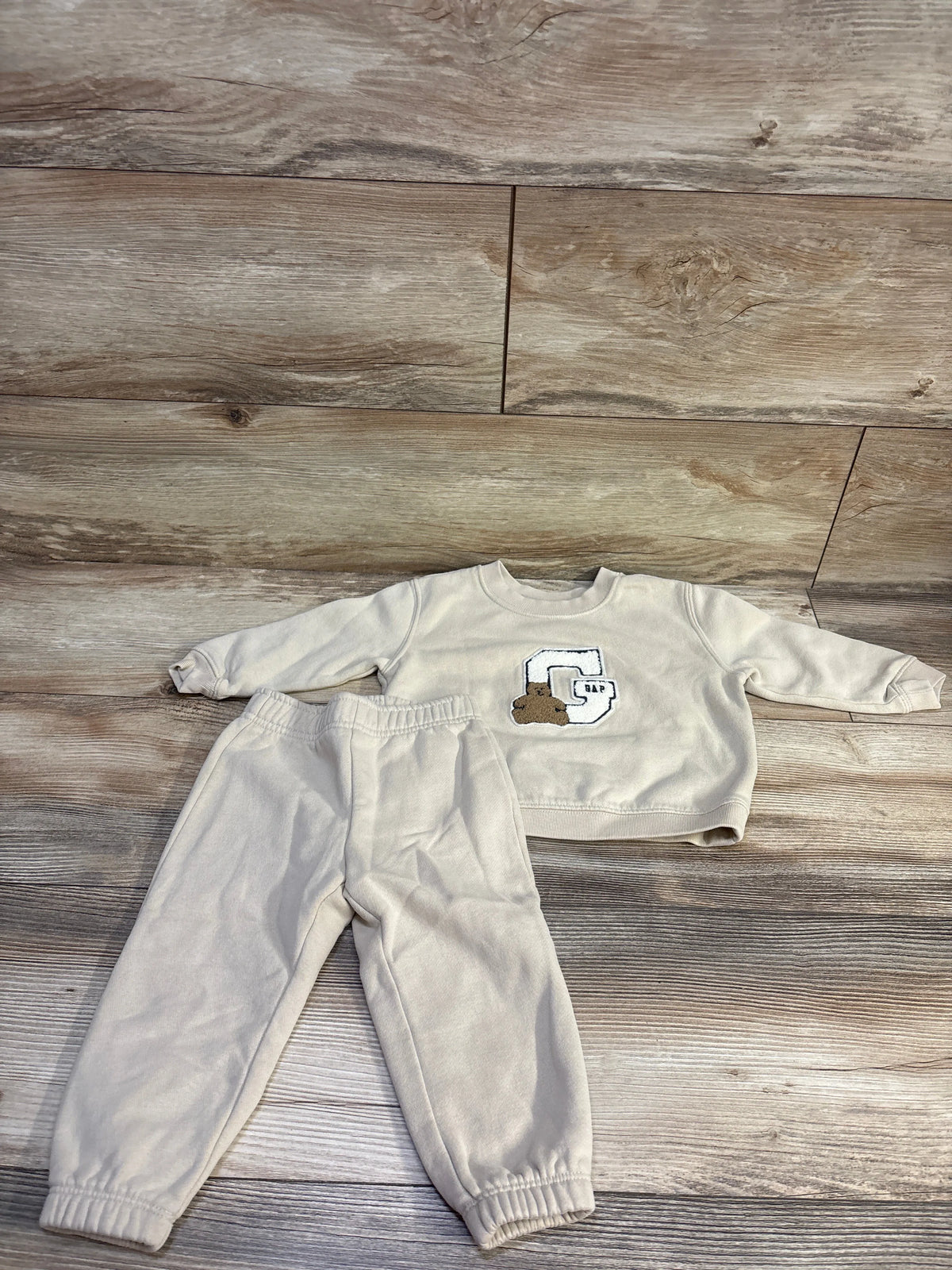 Front view of Baby Gap 2-piece pullover sweatshirt and pants set showcasing beige fabric and Gap bear logo on wood flooring