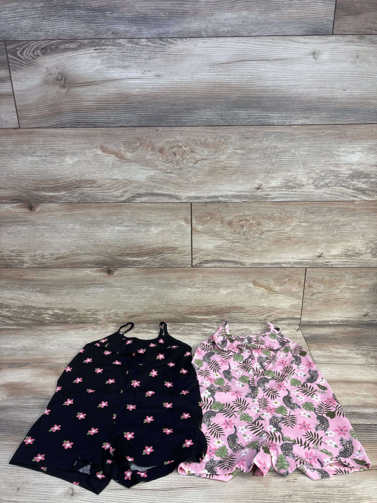 Front view of two Children's Place tank rompers (size 4T) showing black floral romper and pink tropical-leopard romper on wood floor