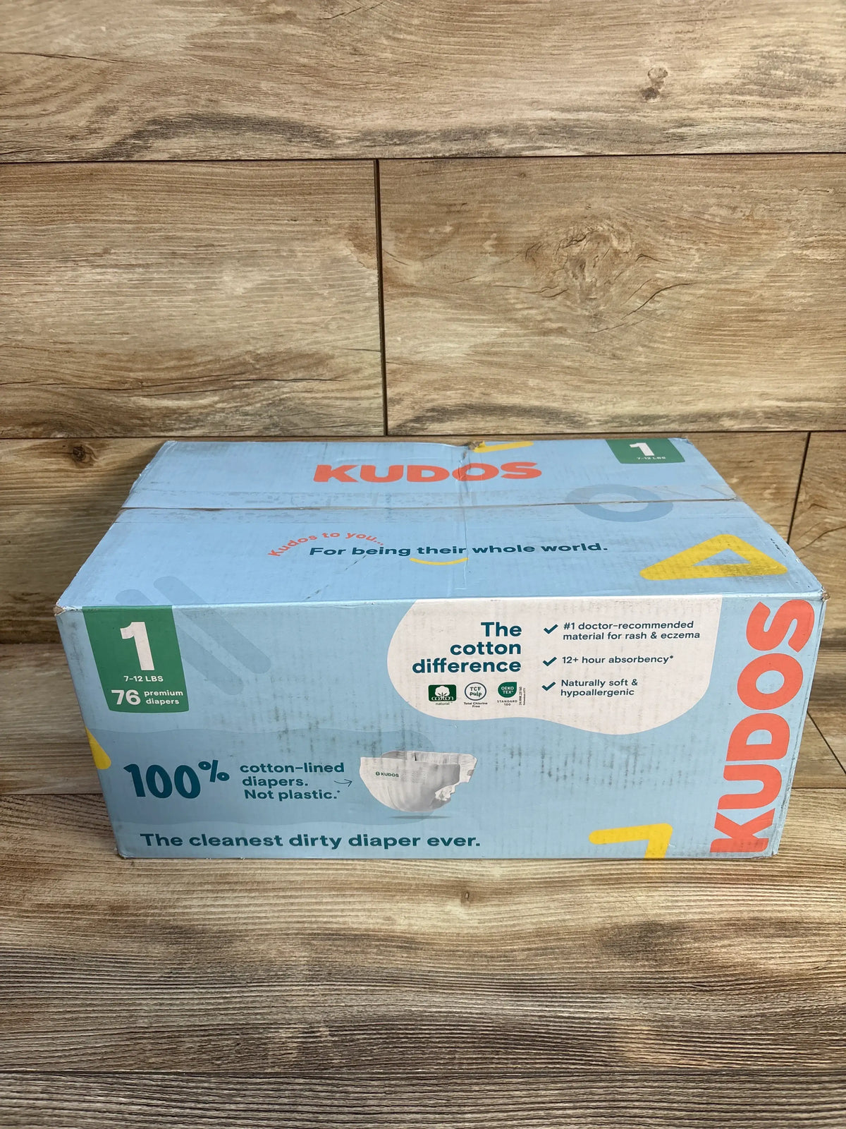 Kudos Size 1 cotton-lined disposable diaper box showing 7 to 12 pound fit and 76 diapers on a wood floor