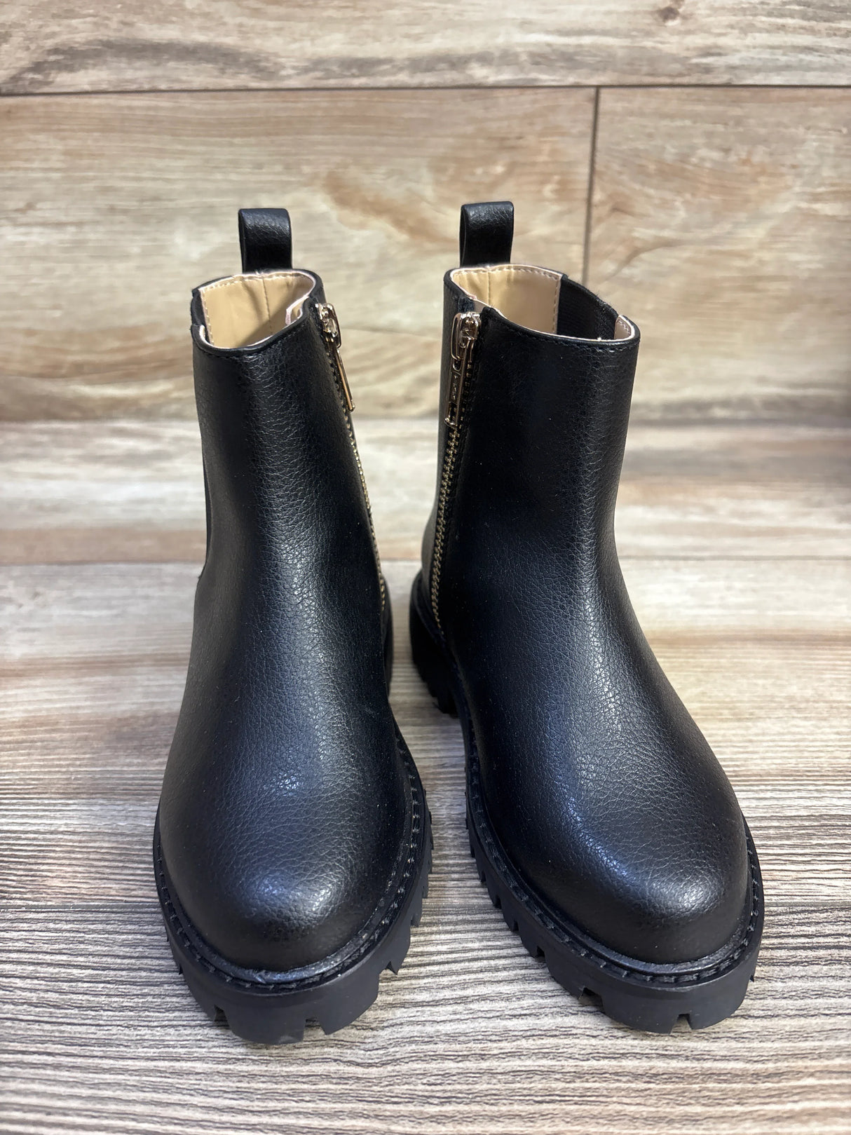 Front view of Janie and Jack Chelsea boots showcasing black faux-leather upper, gold side zippers and chunky lug soles on wood floor