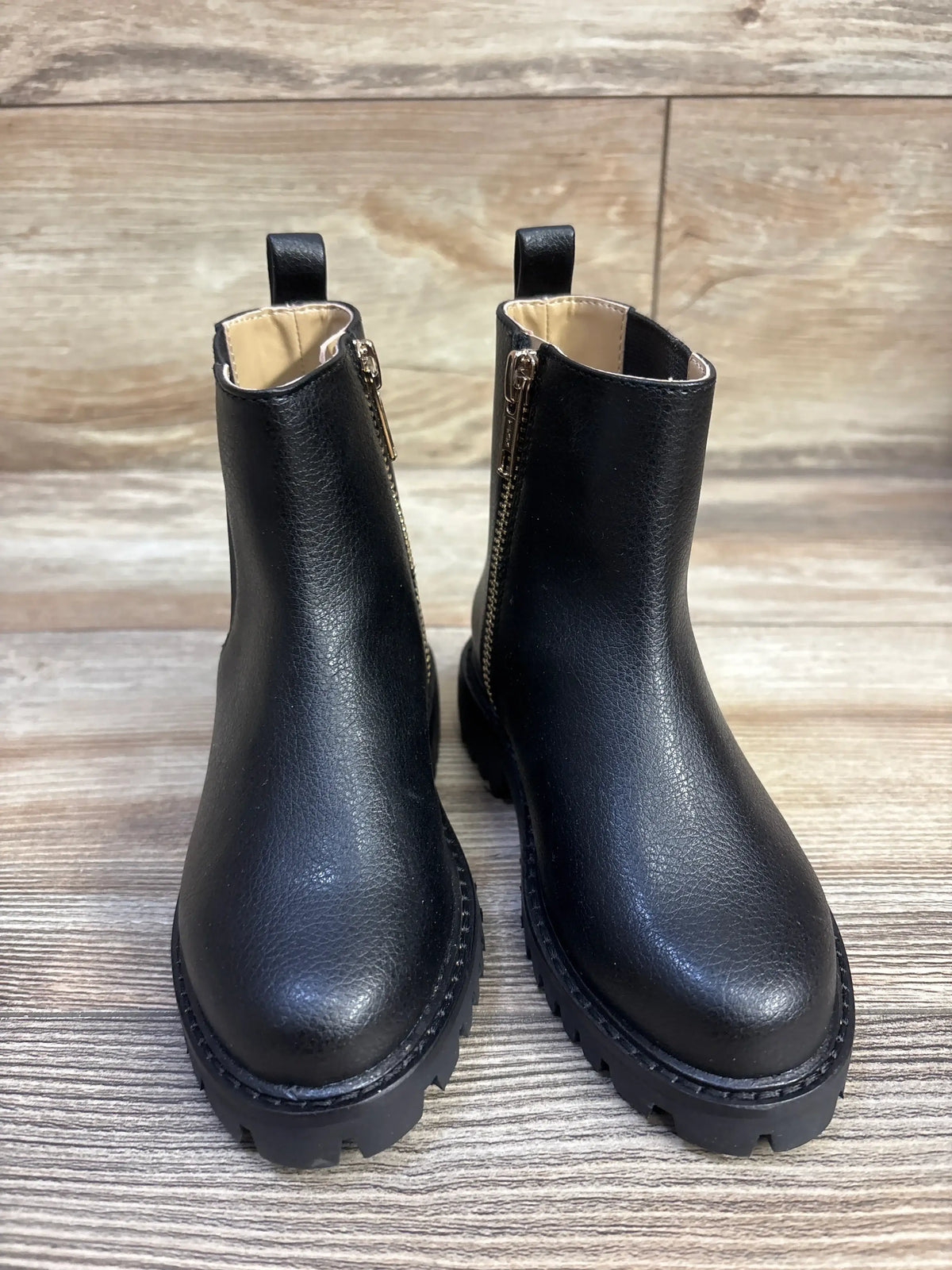 Front view of Janie and Jack Chelsea boots showcasing black faux-leather upper, gold side zippers and chunky lug soles on wood floor