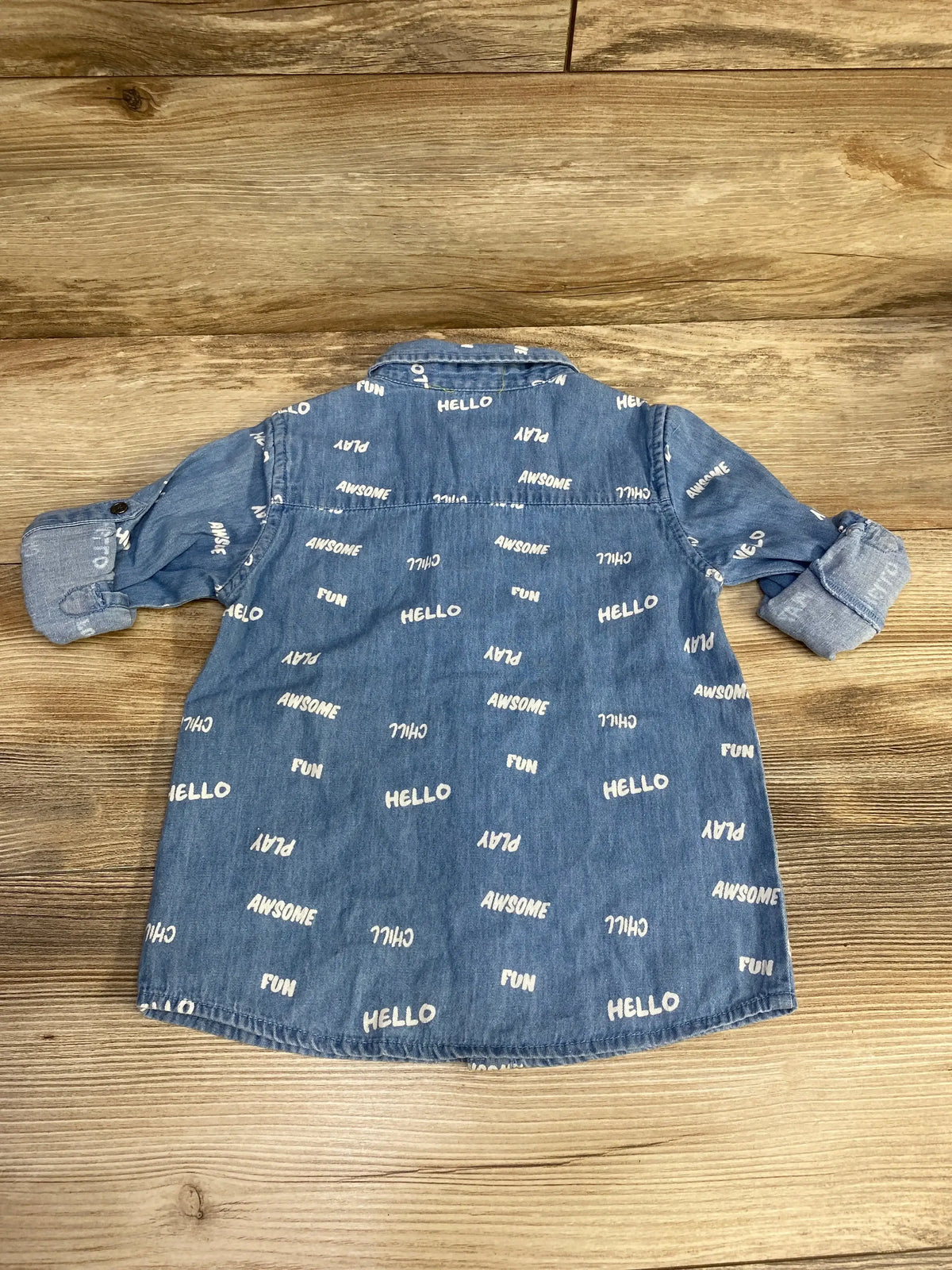 Back view of blue chambray toddler button-up shirt with rolled sleeves and scattered white words HELLO, FUN, PLAY on wooden background