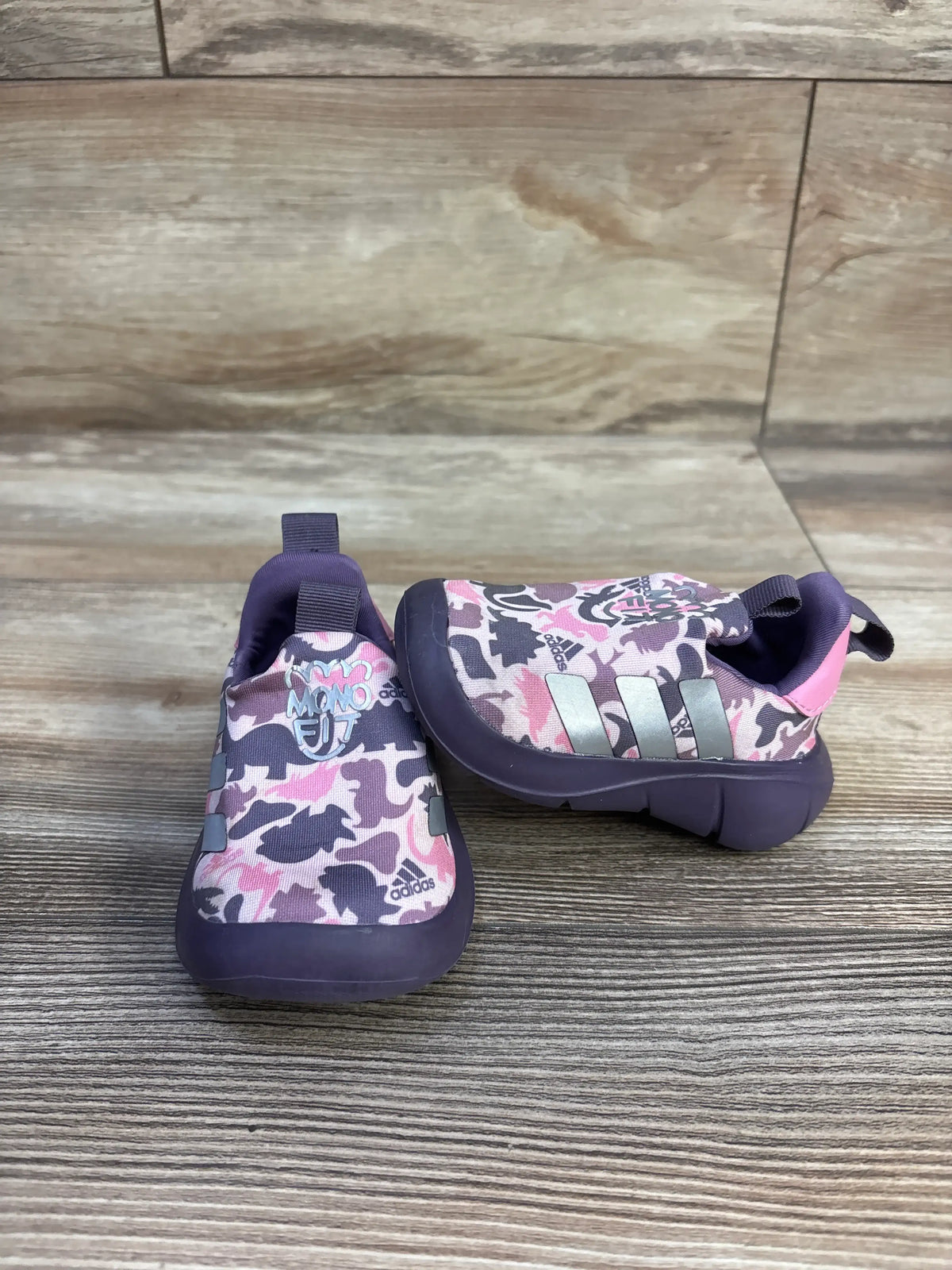 Front and side views of pink Adidas MonoFit slip-on sneakers with camouflage print and pull tabs on a wood-look floor