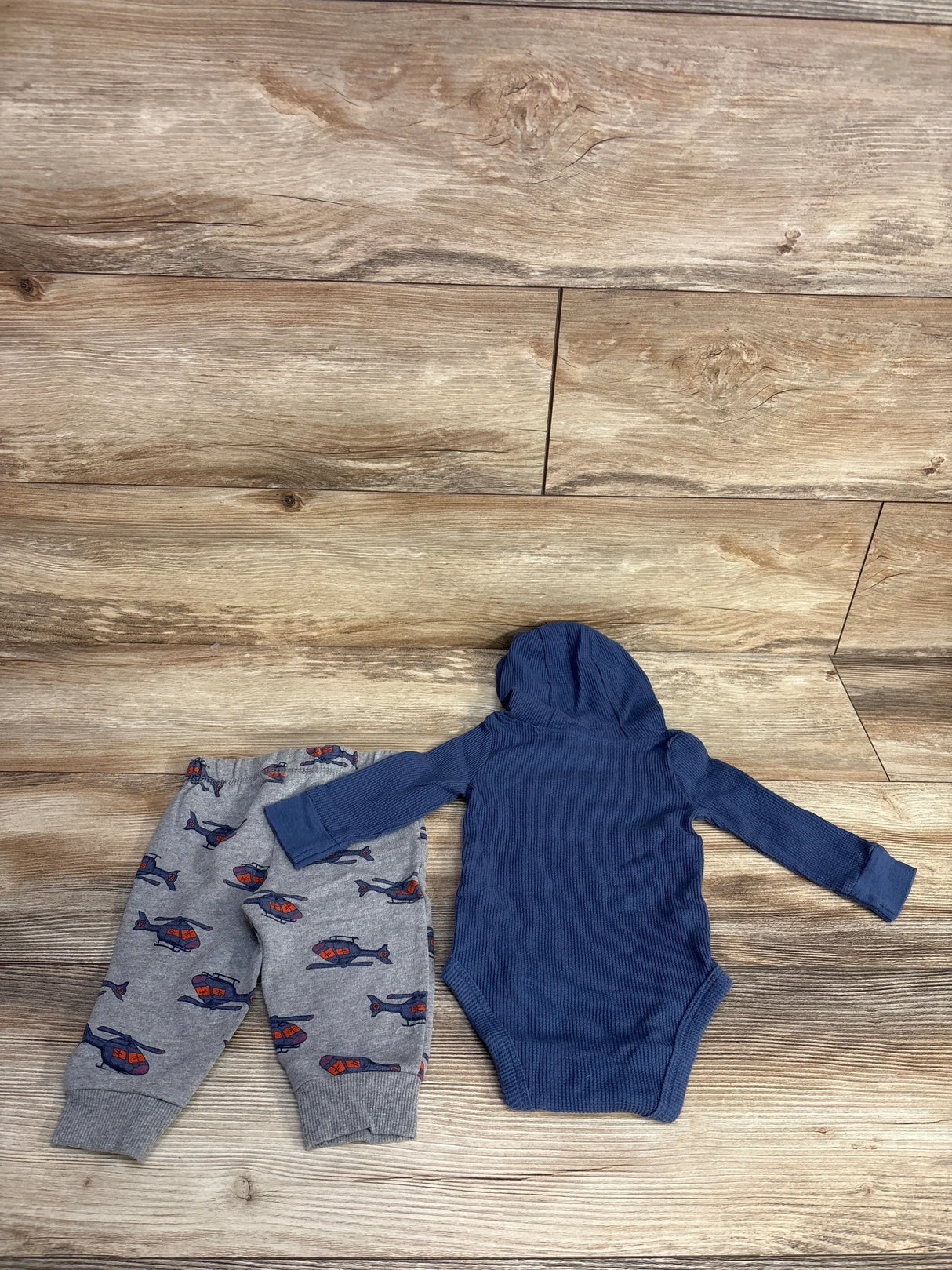 Baby clothing set with grey helicopter print pants and blue hooded long-sleeve onesie on wooden floor