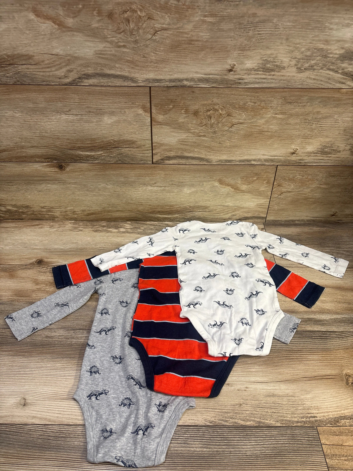 Top view of Carter’s three long-sleeve bodysuits in gray, white dinosaur print, and navy-red stripes on a wood floor