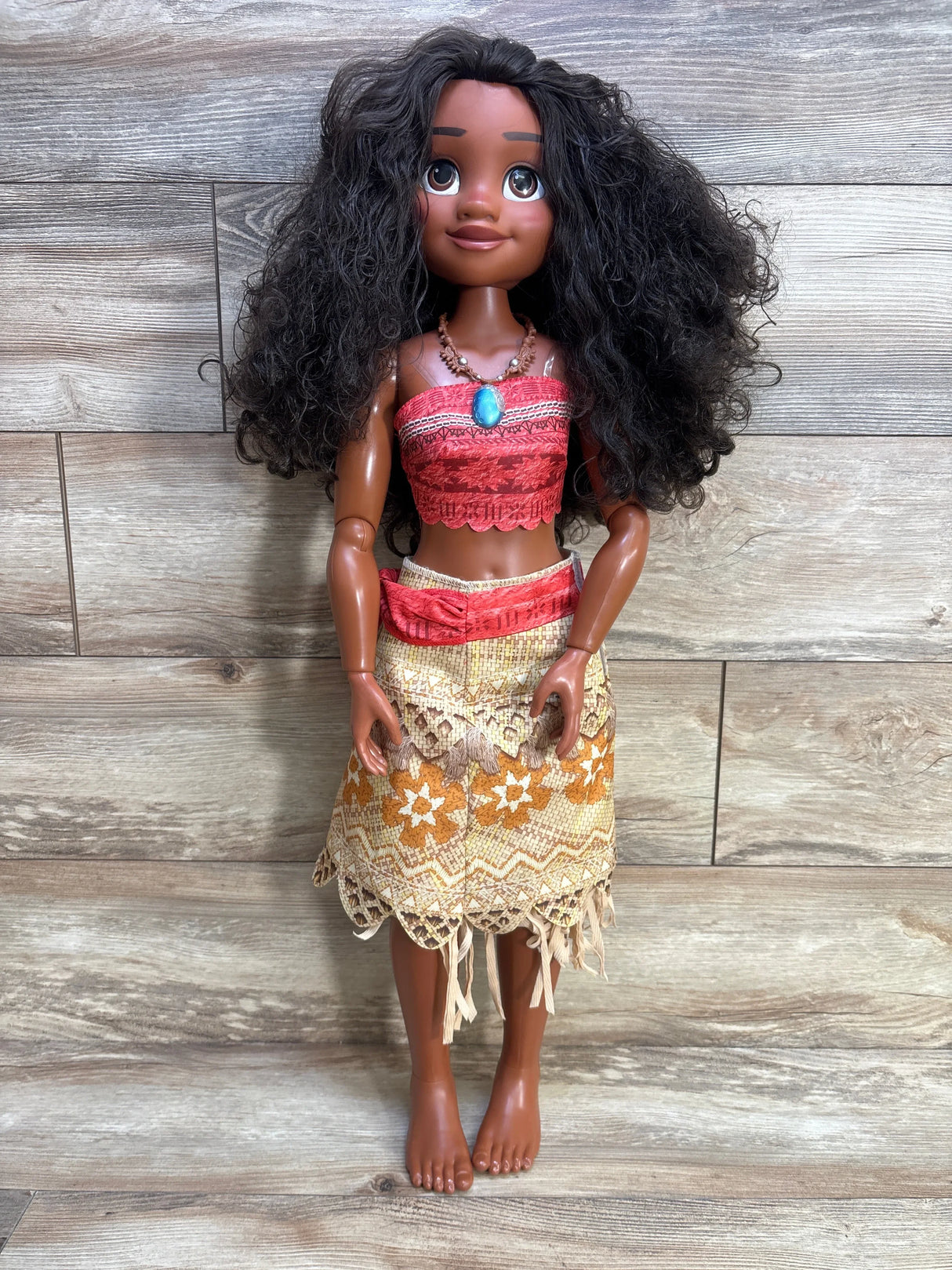 Front view of 32-inch Moana doll with curly hair, red top, woven floral skirt and turquoise pendant against wood background
