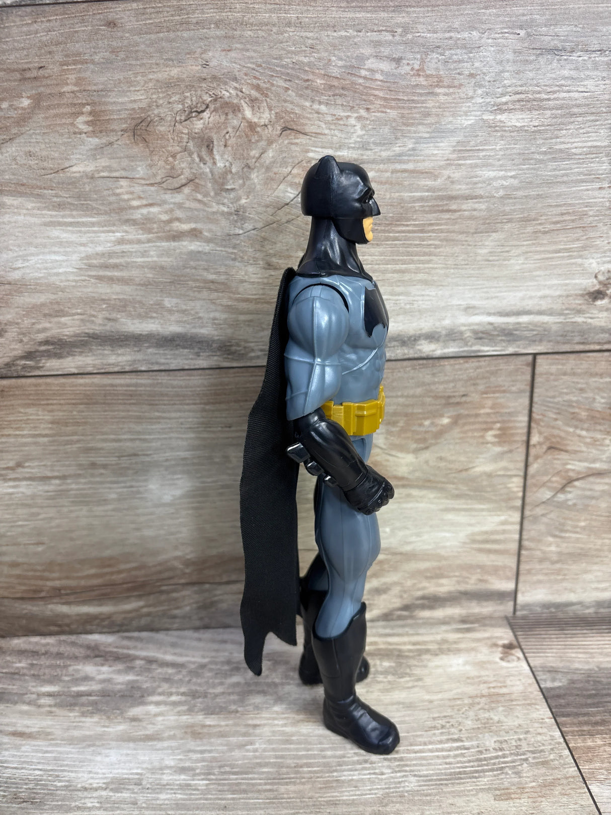 Side view of a Batman action figure with black cape, gray suit, yellow utility belt on wooden background