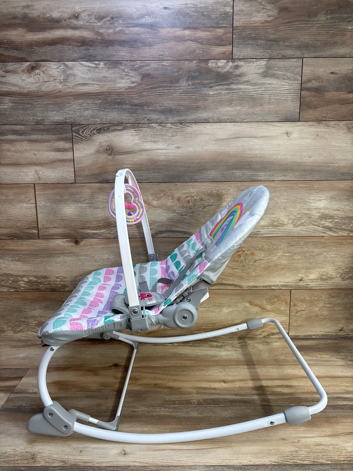 Side view of Bright Starts Rosy Rainbow rocker showing pastel rainbow print and toy bar on wood background