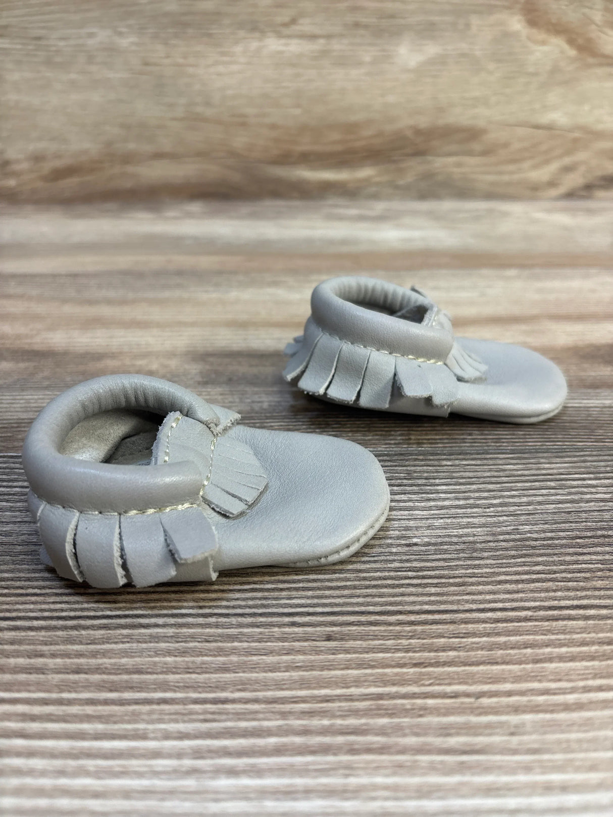 Gray leather baby moccasins with fringe detail on wooden surface