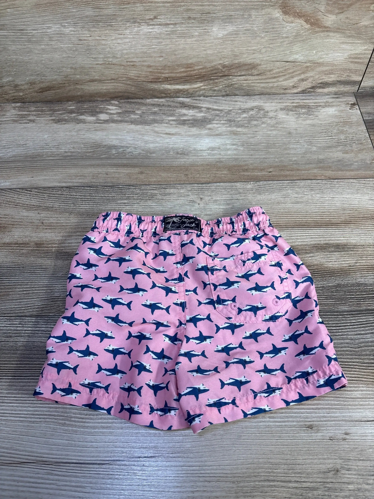 Front view of Janie & Jack swim trunks showing pink with navy shark print, elastic waistband and back pocket on wood surface