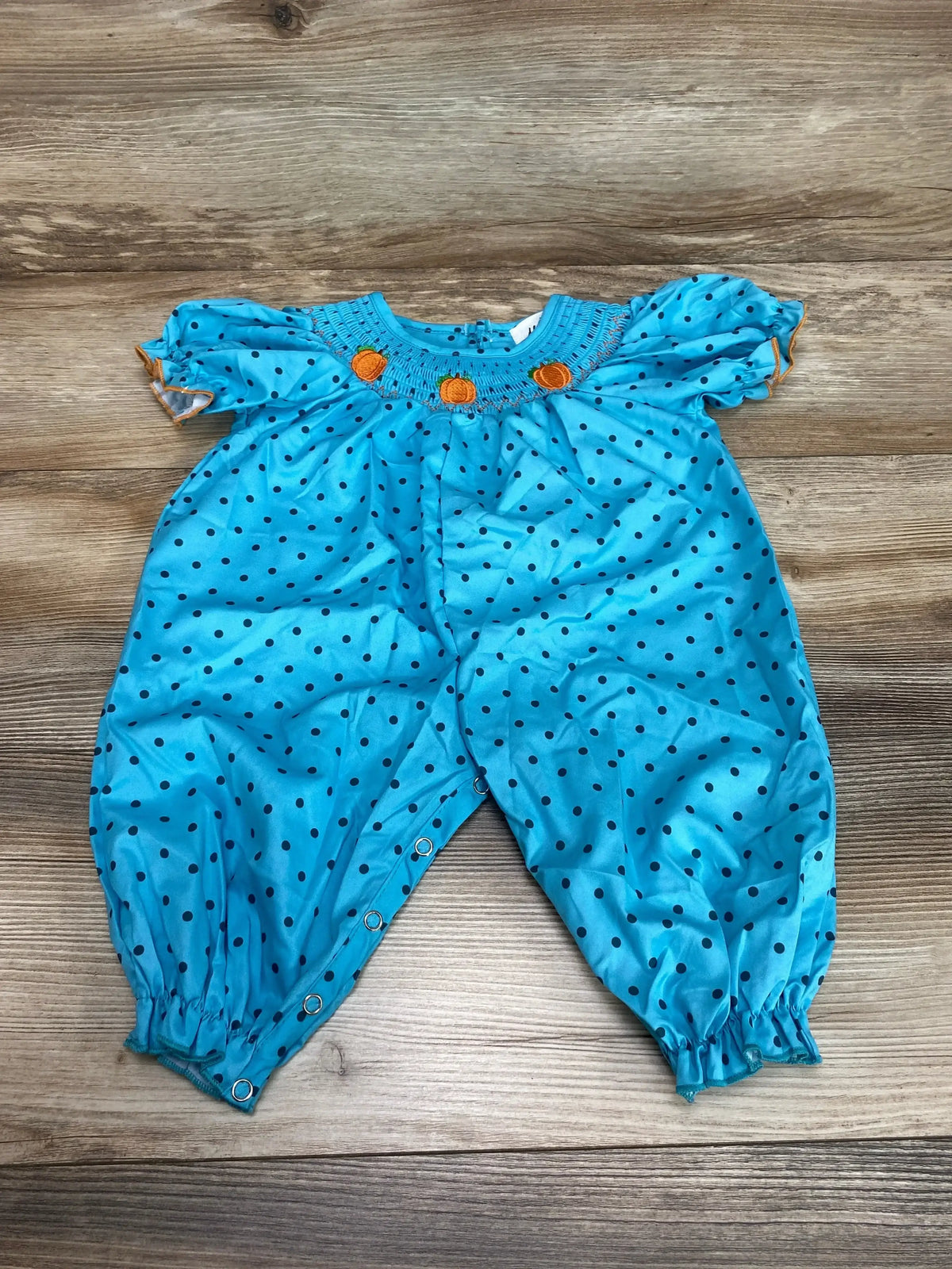 Front view of blue romper with smocked neckline, embroidered pumpkins, polka dots, and cinched sleeves and ankles on wood