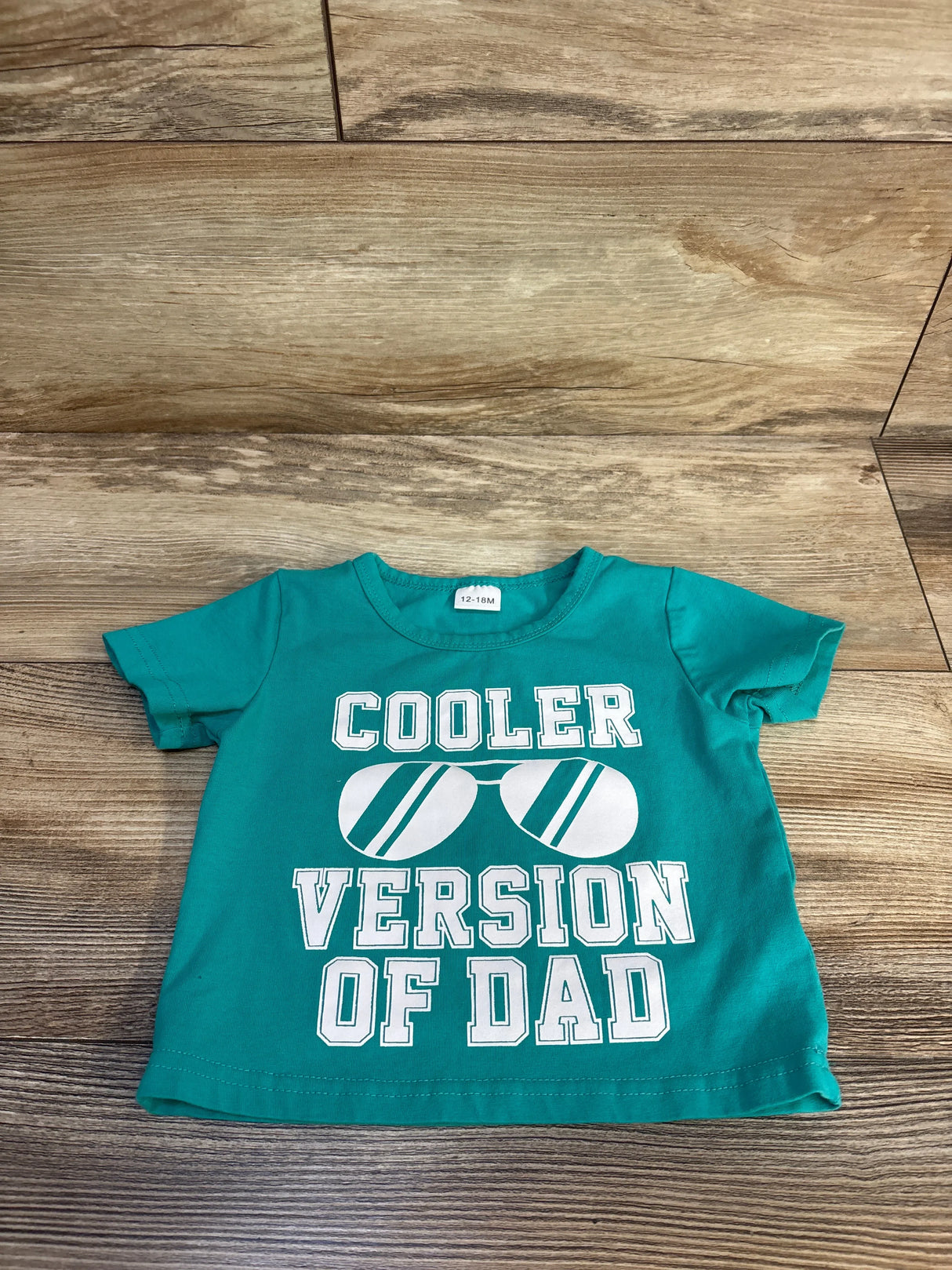 Teal baby tee with 'Cooler Version of Dad' text and sunglasses graphic, 12-18M size on wood floor