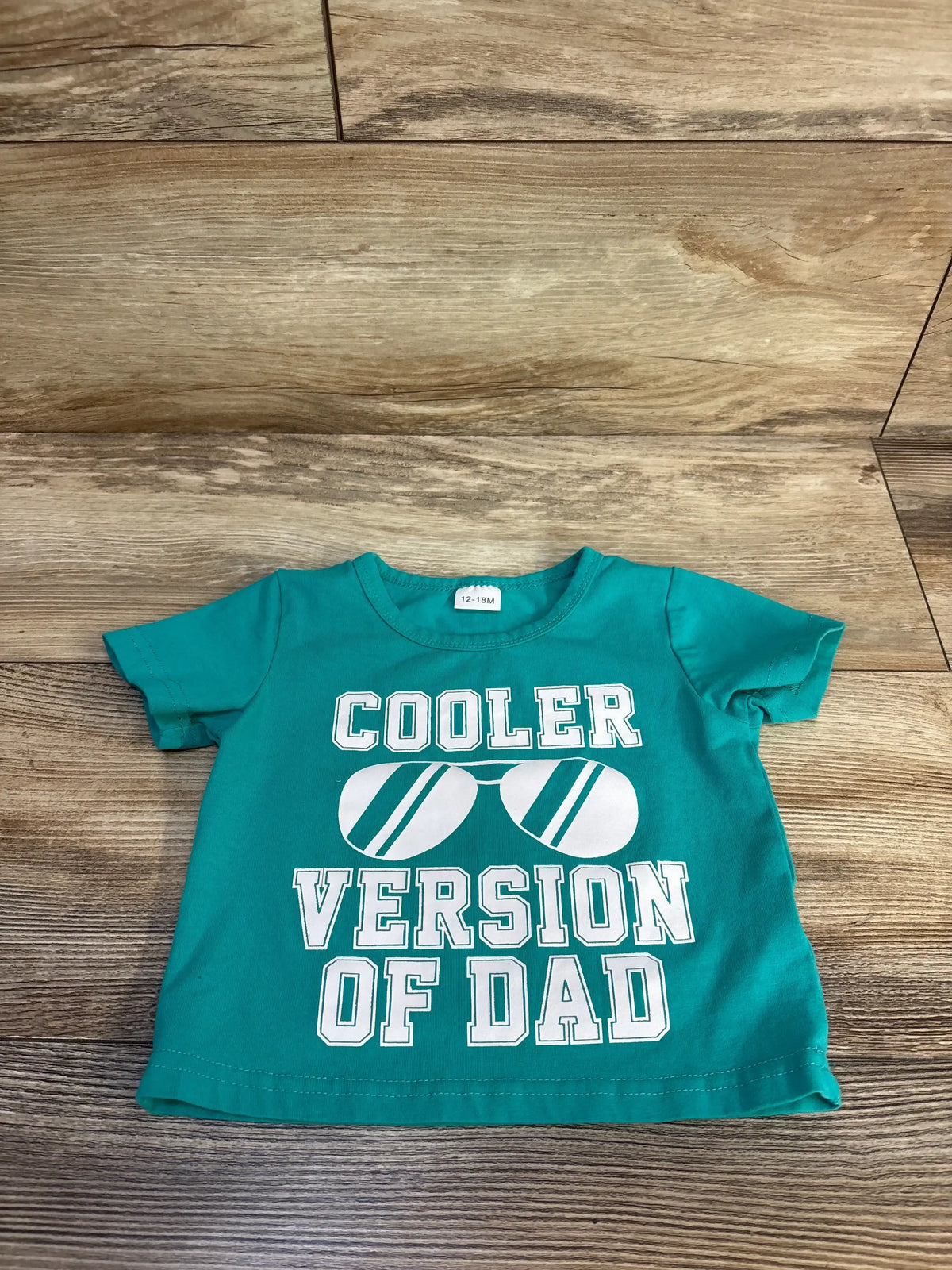 Teal baby tee with 'Cooler Version of Dad' text and sunglasses graphic, 12-18M size on wood floor