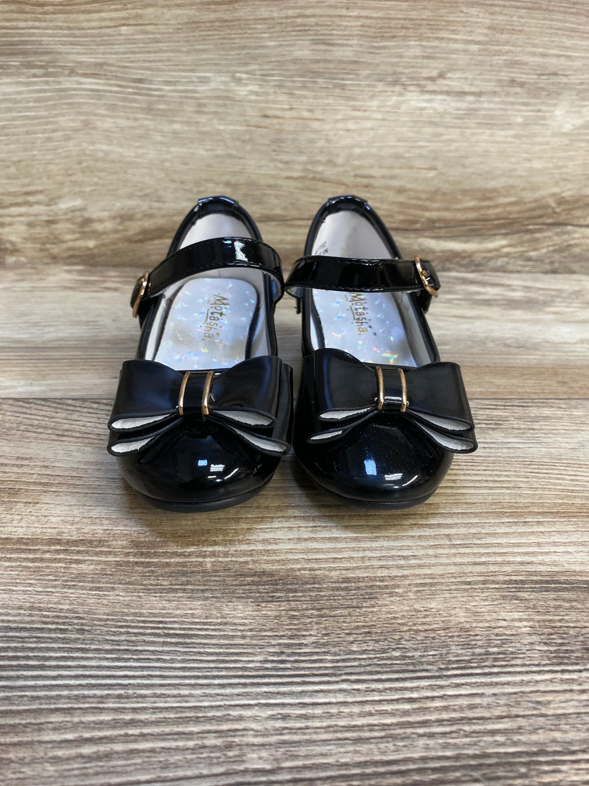 Black patent leather girls' dress shoes with bows and ankle straps on wood floor