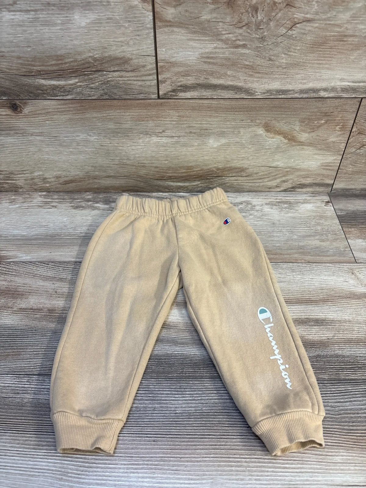 Flat lay of beige Champion joggers showing side script logo and small C logo, elastic waistband and ribbed cuffs on wood background