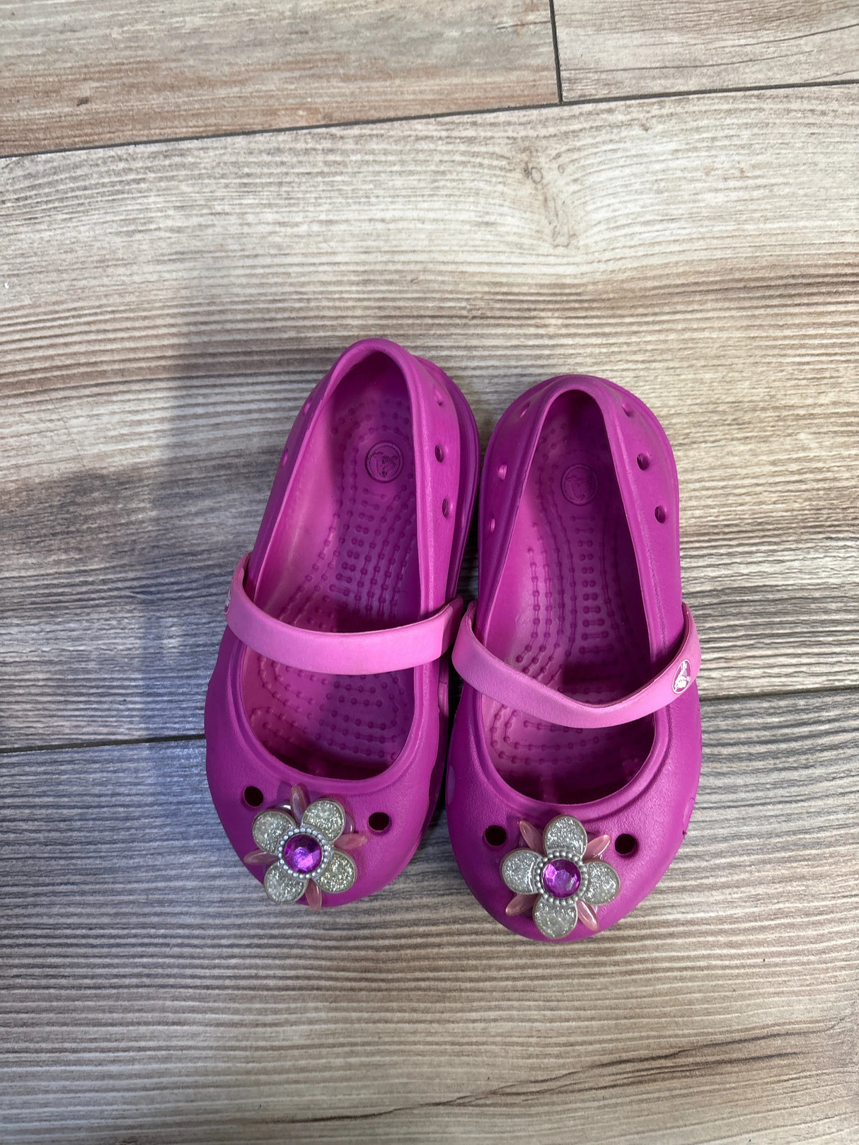 Top-down view of Crocs Keeley Petal Charm flats showcasing pink Mary-Jane strap and flower charm with purple gem on wood floor