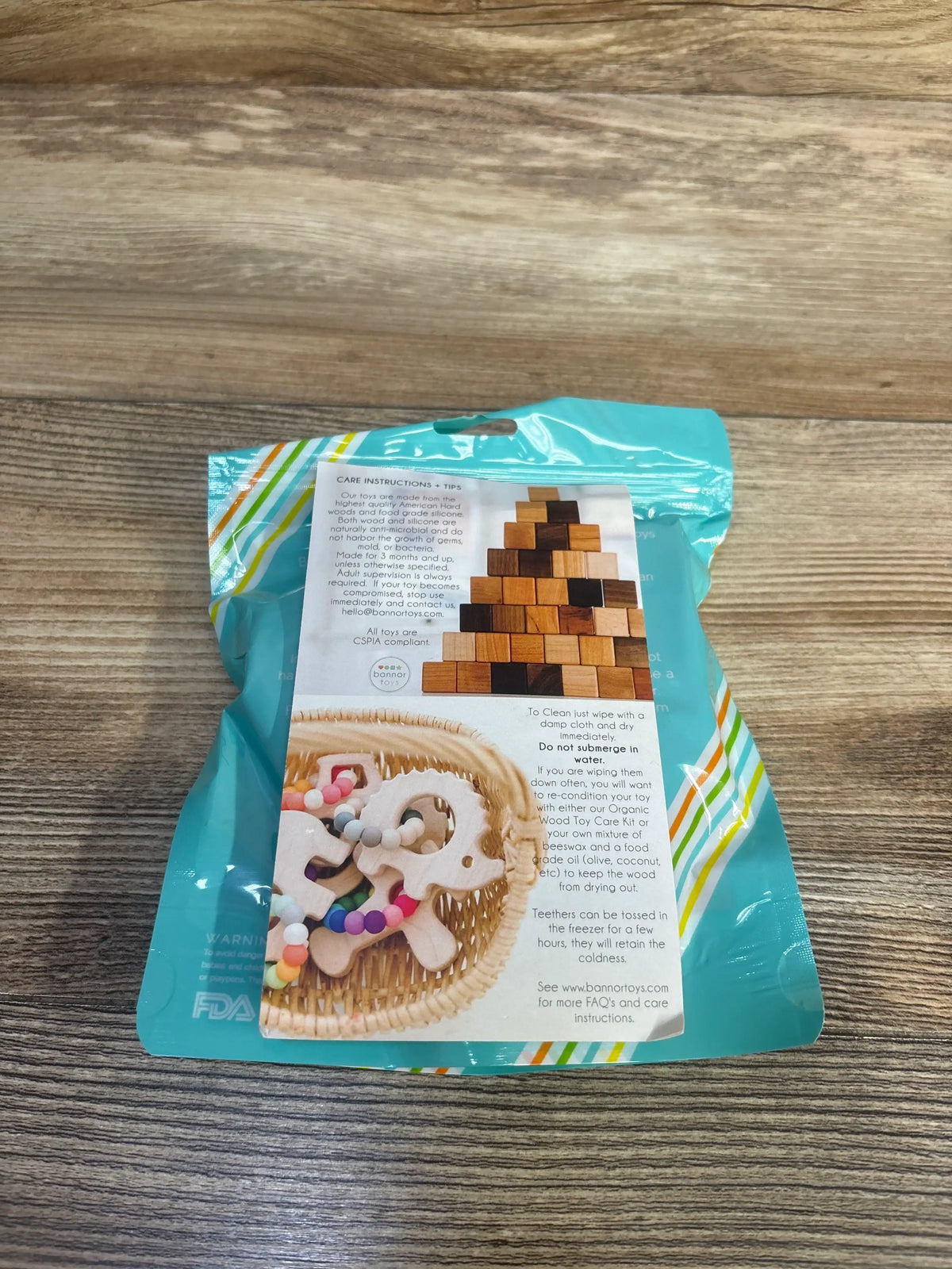 Back view of NEW Bannor Toys Wooden Rattle package showing care instructions and a photo of wooden toys on a wood-grain surface
