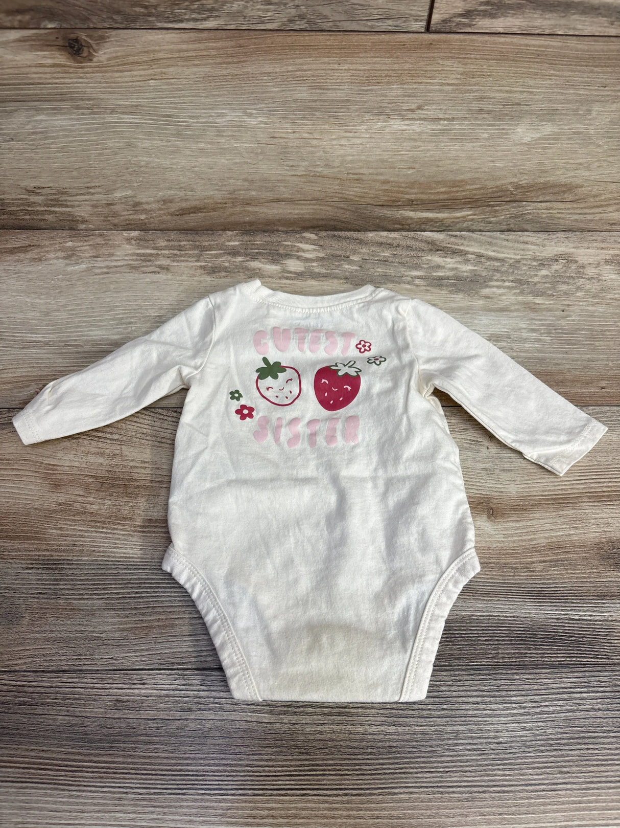 White long-sleeve baby onesie with cute smiling strawberries and pink text reading 'Cutest Sister' on wooden background