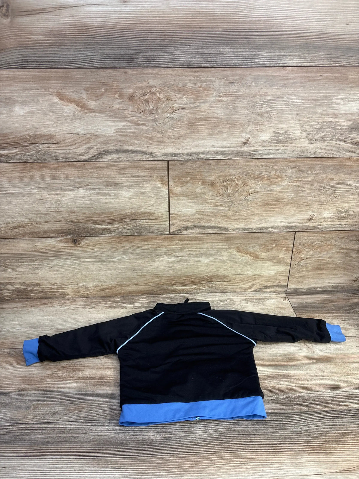 Back view of iPlay zip-up jacket with light blue cuffs, hem, and shoulder piping; laid flat on wood, 18m