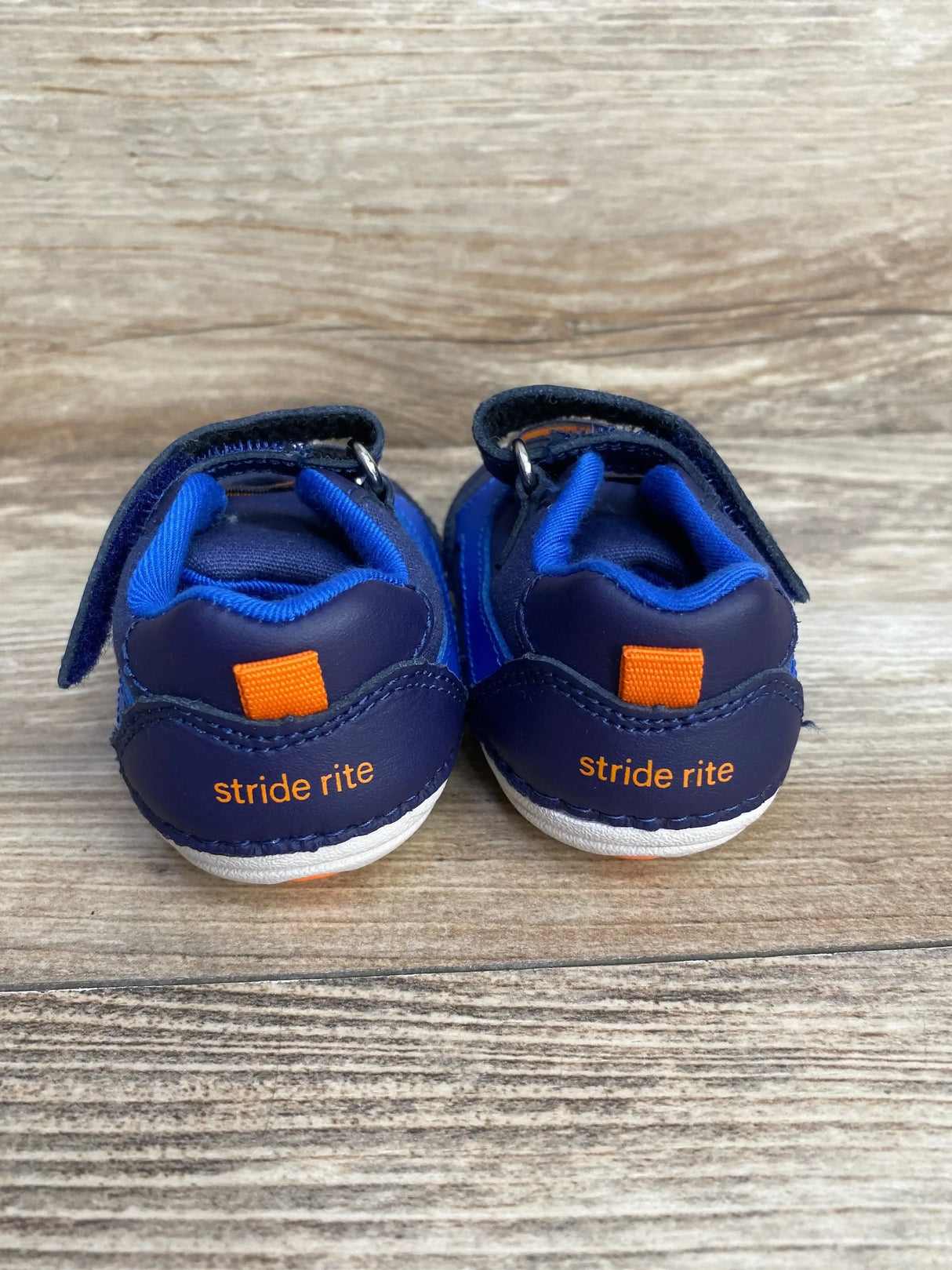 Rear view of navy blue Stride Rite Soft Motion Kylin baby sneakers showing velcro straps, orange heel tabs and stride rite logo on wooden floor